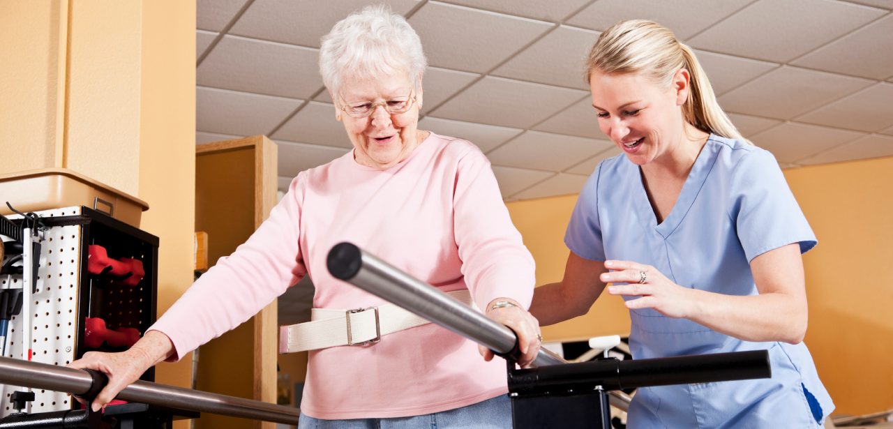 Senior woman (80s) on parallel bars doing gait training with physical therapist (30s).