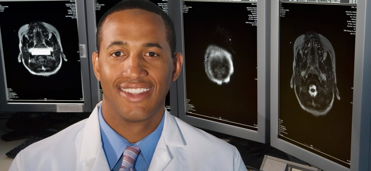 Detroit, Michigan, USA --- Man smiling in front of brain scans --- Image by © Paul Burns/Blend Images/Corbis