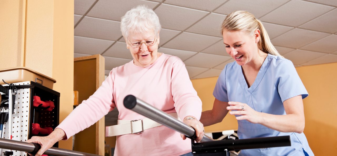 Senior woman (80s) on parallel bars doing gait training with physical therapist (30s).