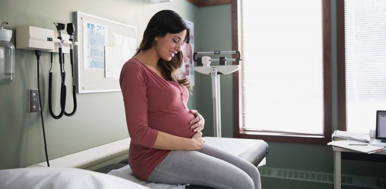 10 Dec 2014 --- Smiling pregnant woman holding stomach in examination room --- Image by © Hero Images Inc./Hero Images Inc./Corbis