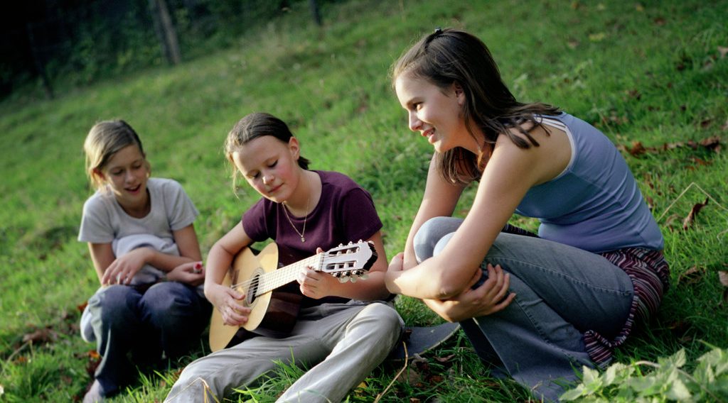 Three Girls Sitting with Guitar in Meadow --- Image by © Mika/Corbis