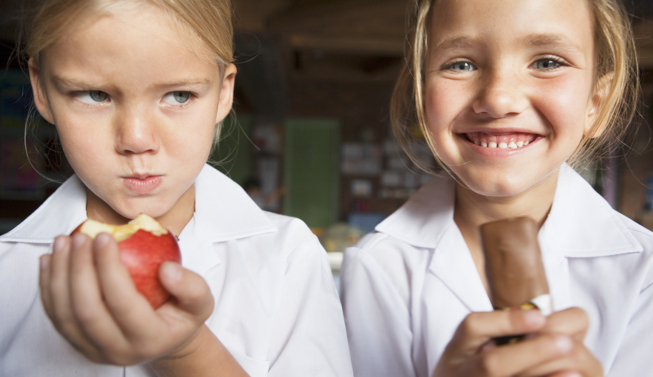 Angry Sibling Holding an Apple While Her Twin Eats Chocolate --- Image by © Roy McMahon/Corbis