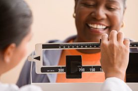 overweight African American woman standing on scale at doctor's office, smiling, happy. 
Source: 85103,PWS_BLP0020114